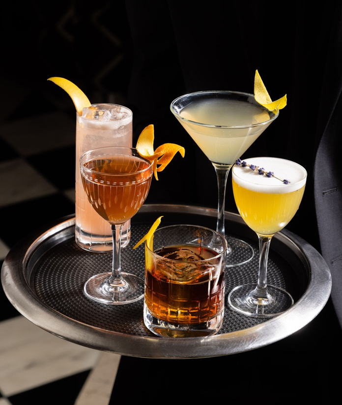 Five colorful cocktails with citrus garnishes arranged on a round black serving tray against a dark background