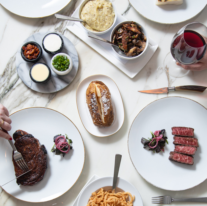 Modern restaurant with green booth seating, chandeliers, wooden shelves, and neatly arranged tables with glassware.
