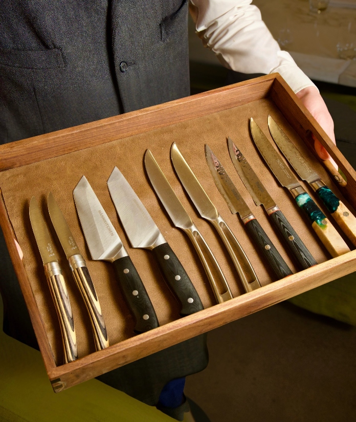 A server holding a wooden tray lined with assorted steak knives, including several styles of handles and blade shapes, presented for a guest to choose from.