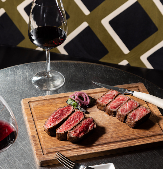Sliced medium-rare steak on wooden cutting board with pickled onions, served alongside two glasses of red wine