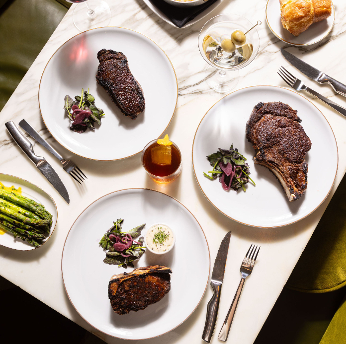 Three plates of grilled steak with salad garnish on white marble table with wine glasses and asparagus side dish