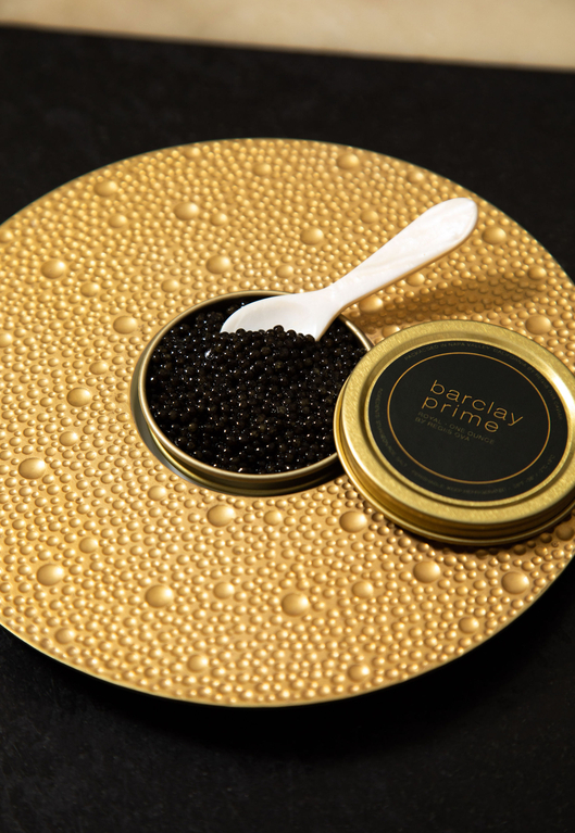 Caviar in gold tin with mother-of-pearl spoon on golden beaded plate against black background