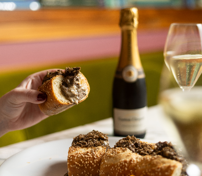A hand holding a Cheesesteak topped with shaved truffles, served alongside a chilled bottle of champagne.