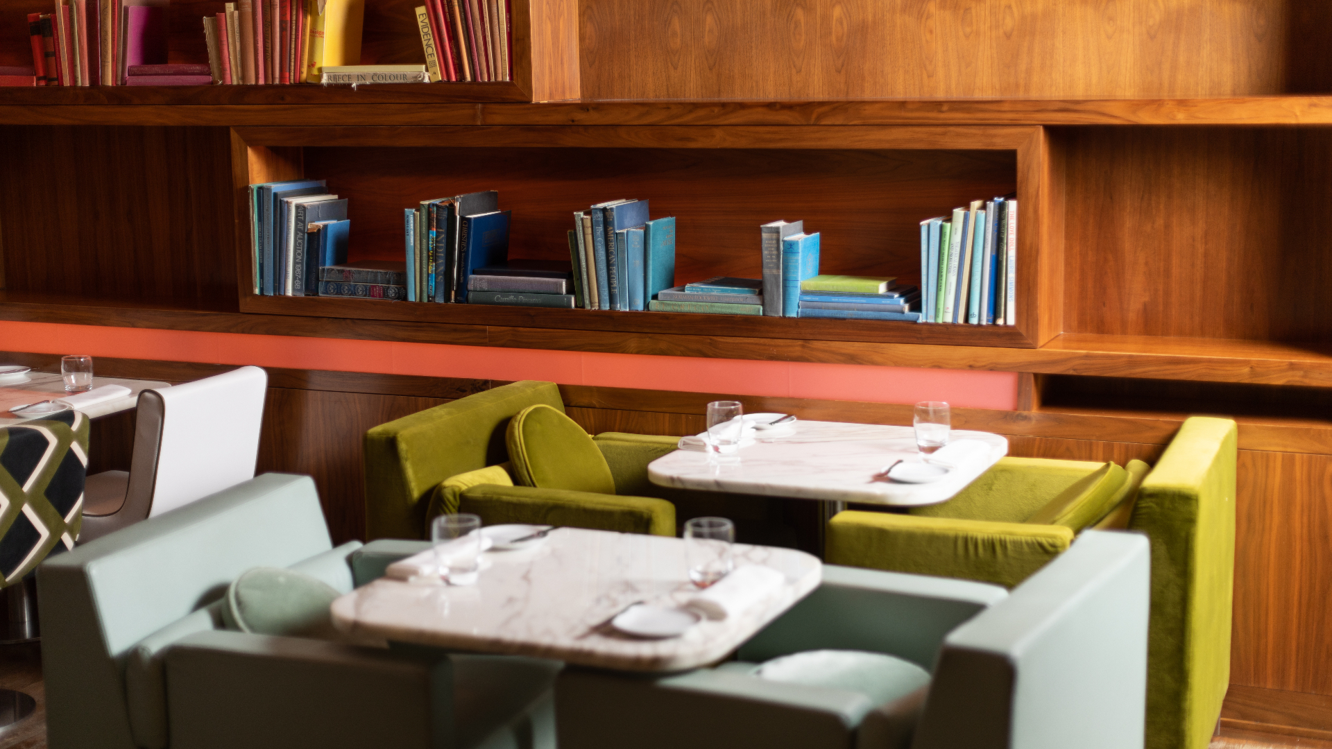 Cozy restaurant interior with green velvet chairs, marble tables, and wooden shelves filled with books.
