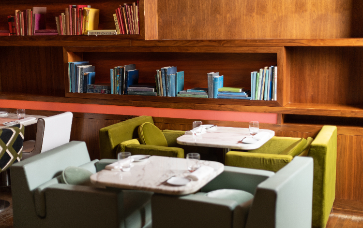 Restaurant interior with wooden shelves filled with books, tables with white tops and green booth seating.