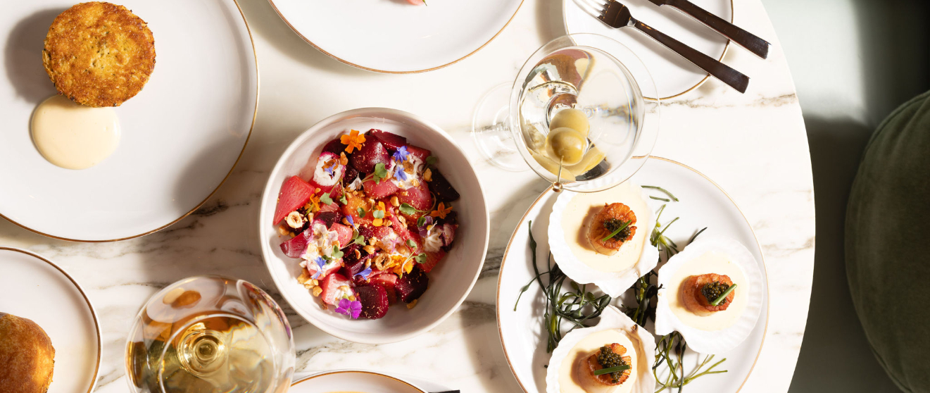 Overhead view of elegant plated dishes including a colorful beet salad, scallops, fried croquettes, and white wine glasses